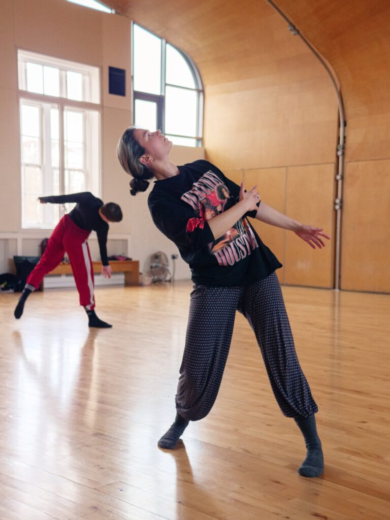 An image of two dancers in a dance studio. Both dancers are standing and bending sideways.