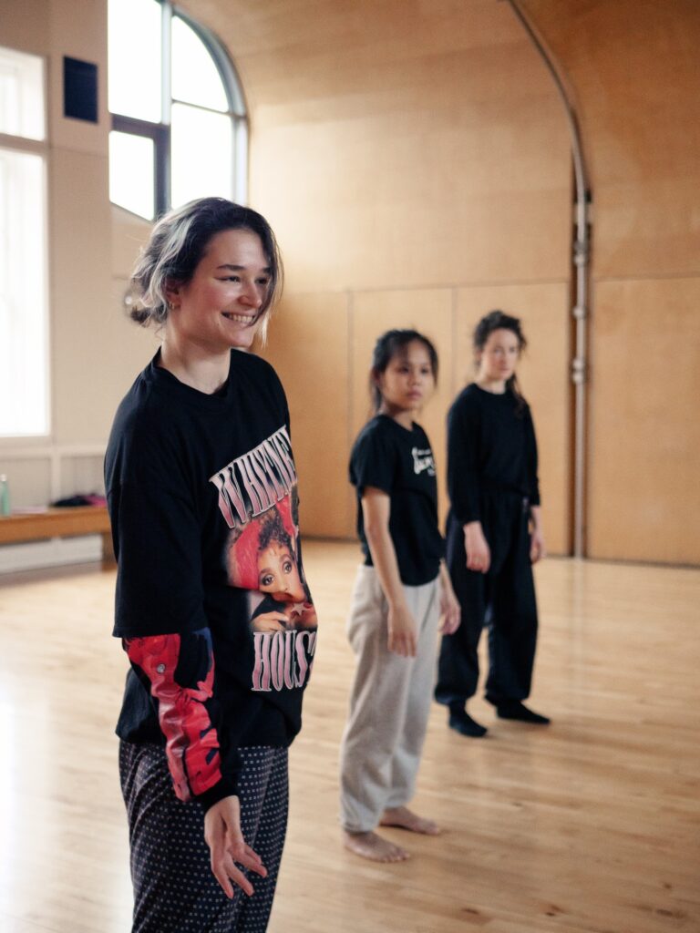 An image of three dancers standing in a dance studio smiling and looking out.