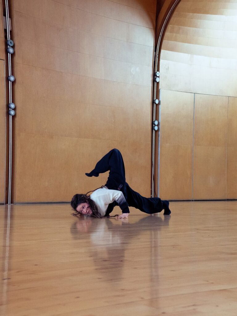 An image of a dancer in a dance studio. They are on the floor on their chest bending one leg up in the air.