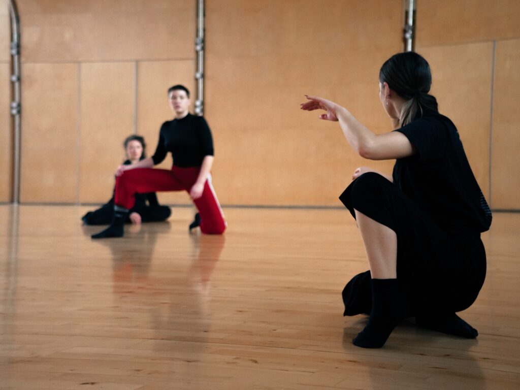 An image of three dancers sitting on the floor. The one in the front is facing the back of the studio and two other dancers are in the background facing the dancer in the front.