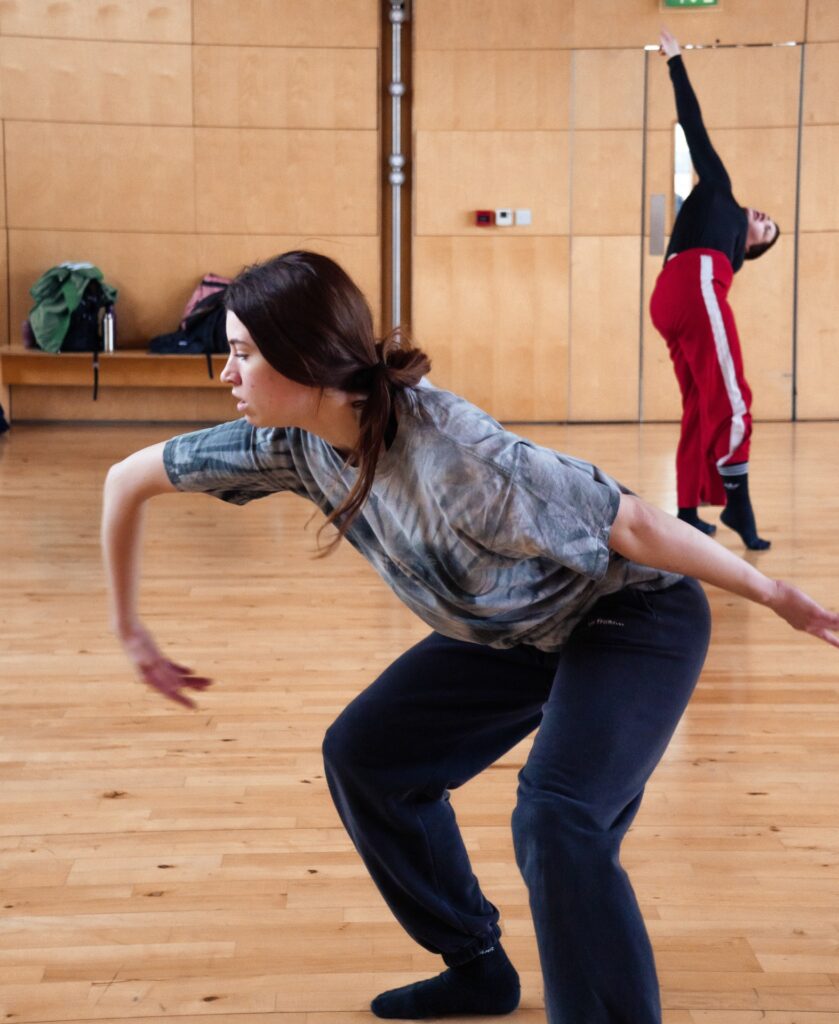 An image of two dancers dancing in a dance studio. The dancer in the front is bending legs and leaning towards front with their chest, one arm curved and one arm extended. The dancer in the background is standing and reaching one arm towards the ceiling.