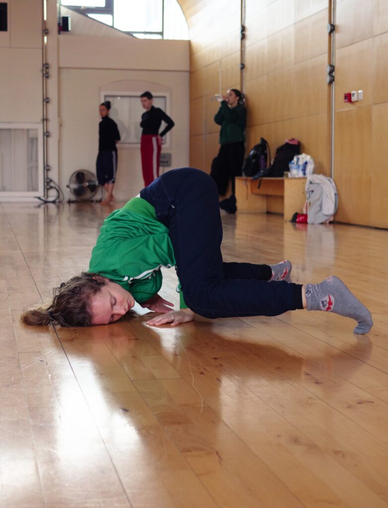 An image of a dancer balancing on their hand, feet and head on the floor. Three dancers are standing in the background, one of them is drinking water..