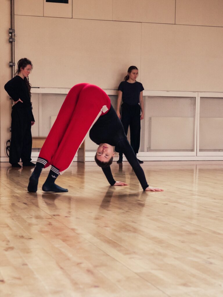 An image of a dancer upside down balancing on their feet and arms. There are two dancers standing in the background.