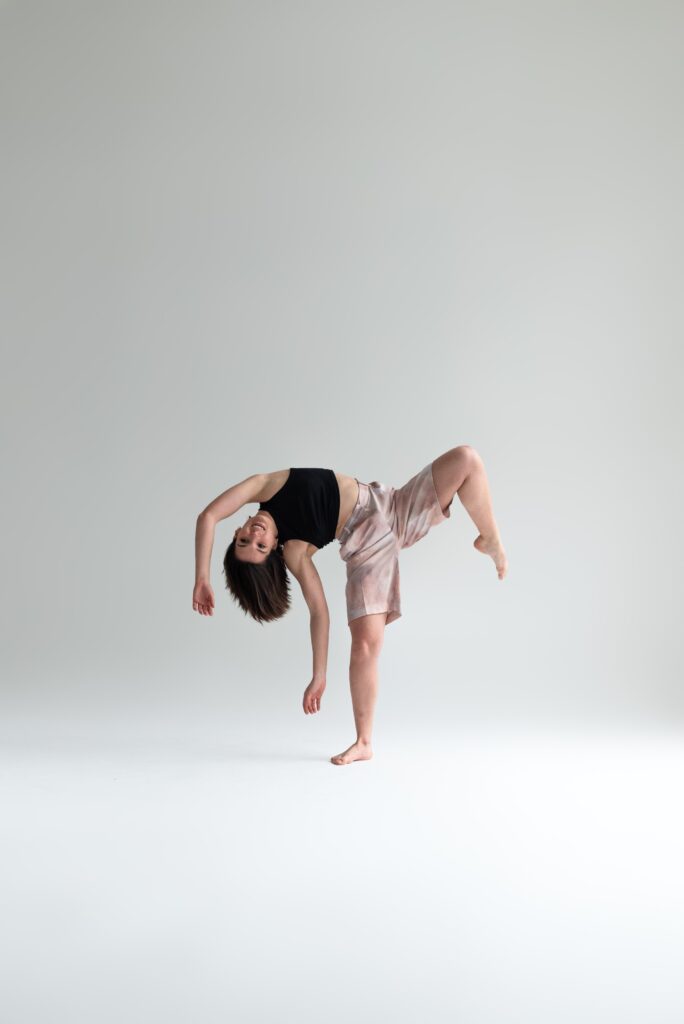 Laure wears a black top and white shorts. She is balancing on one leg, the other leg is up and her chest arms and head are falling on the other side.