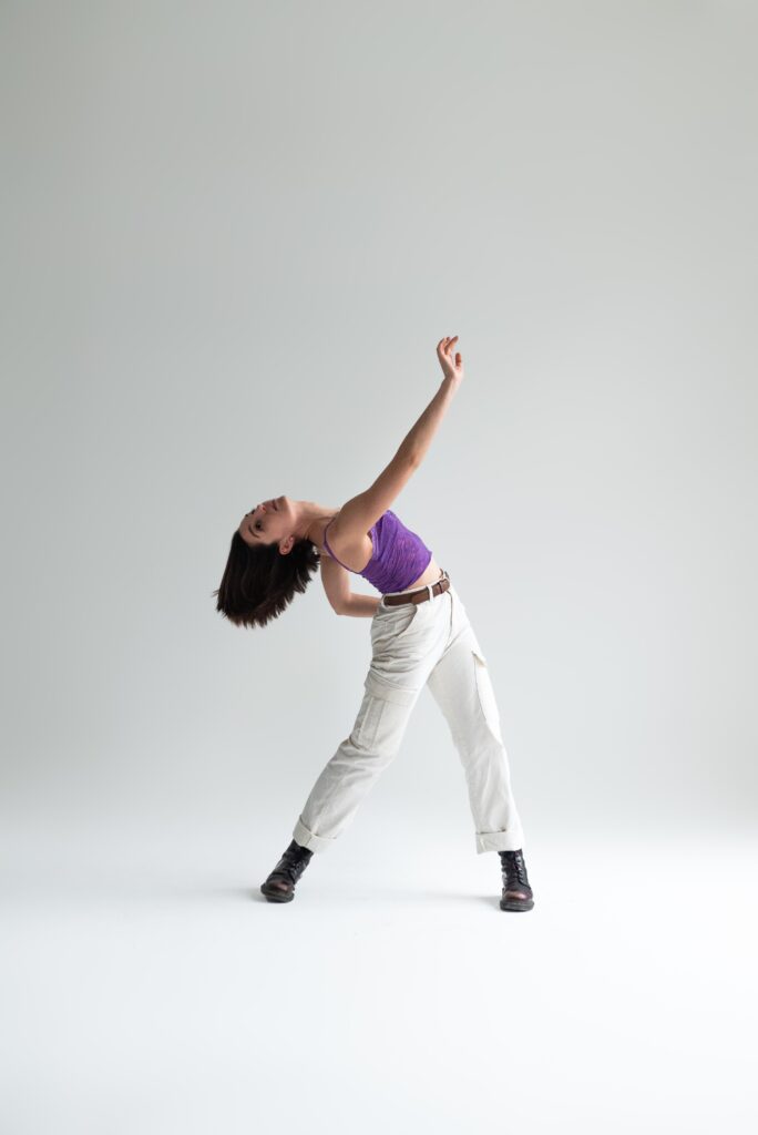 Laure is wearing a purple topand white trousers. Her head is falling back in a back bend and one arm is reaching up.